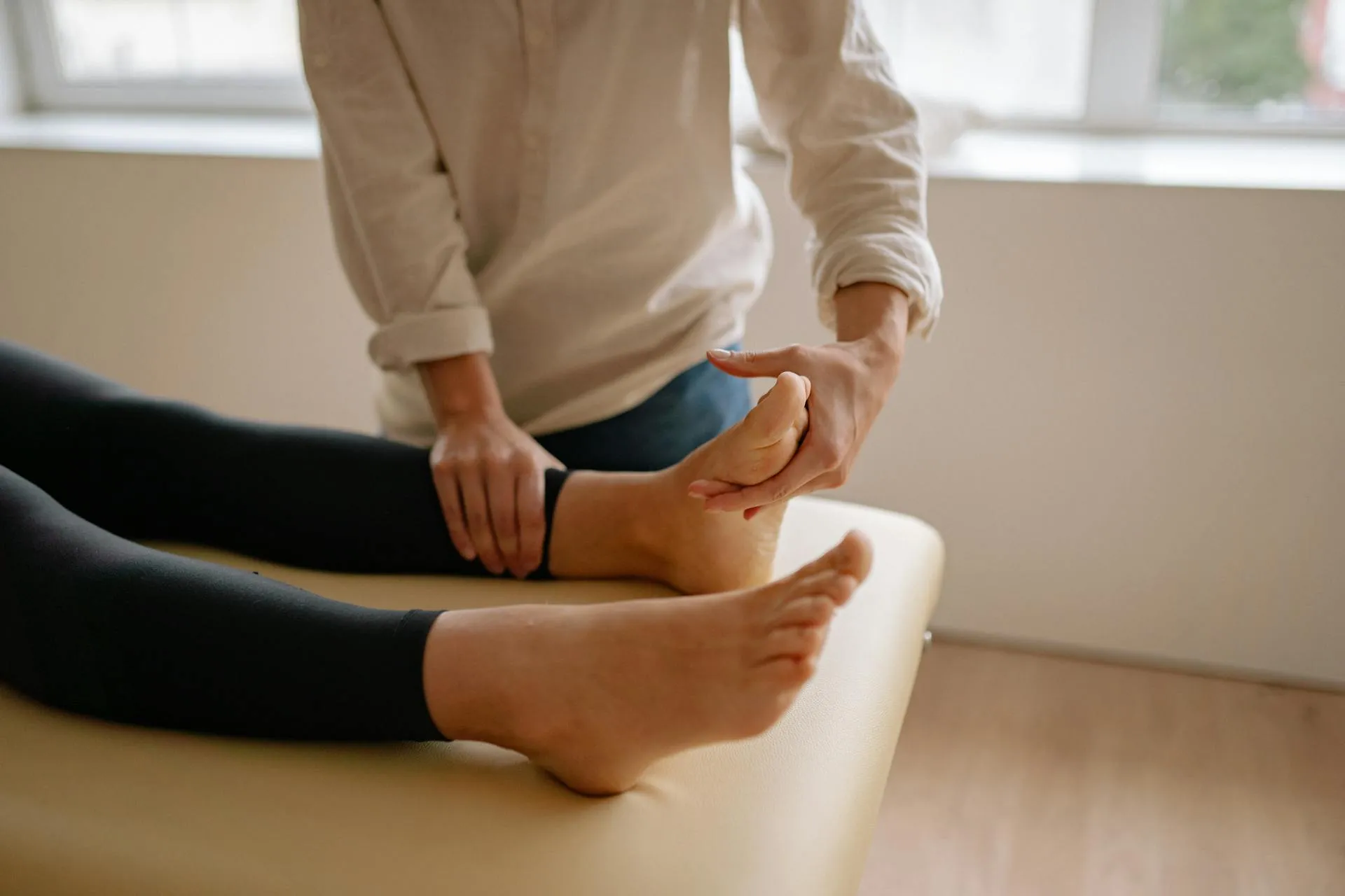 A close-up view of a foot massage session. A person in a light-colored shirt is gently holding and manipulating the ankle of another person, who is lying on a massage table. The person receiving the massage is wearing black leggings, and their foot is bare. The background features a window with soft natural light coming in, creating a calm and soothing atmosphere.