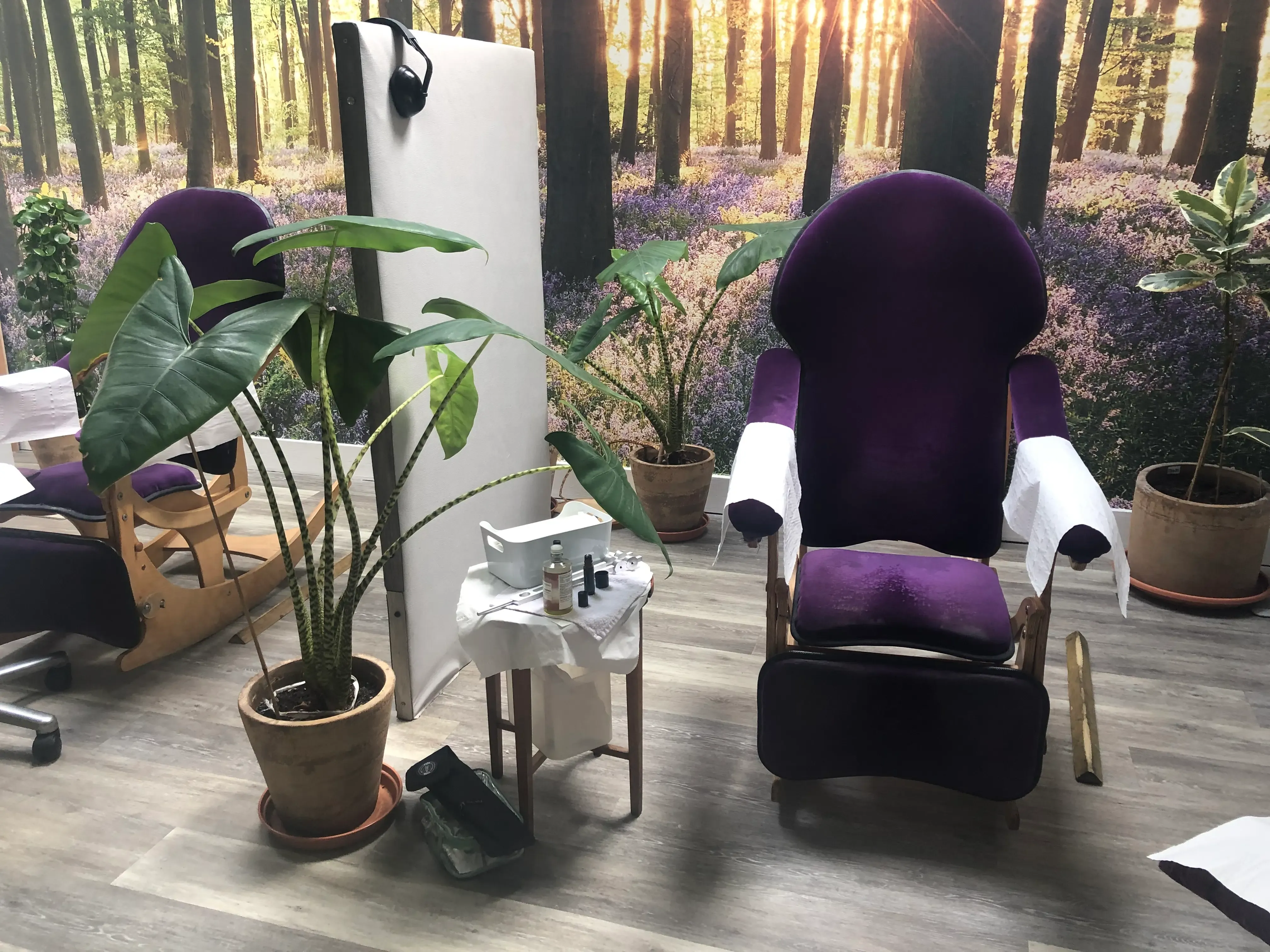 A cozy spa treatment room featuring a plush purple treatment chair with white towels draped over the arms. In the background, a large wall mural depicts a serene forest scene with sunlight filtering through trees. There are several potted plants, including a tall leafy plant in the foreground, and a small table with spa supplies like nail polish and a bottle. A partition stands to the left, and the flooring is light-colored wood.