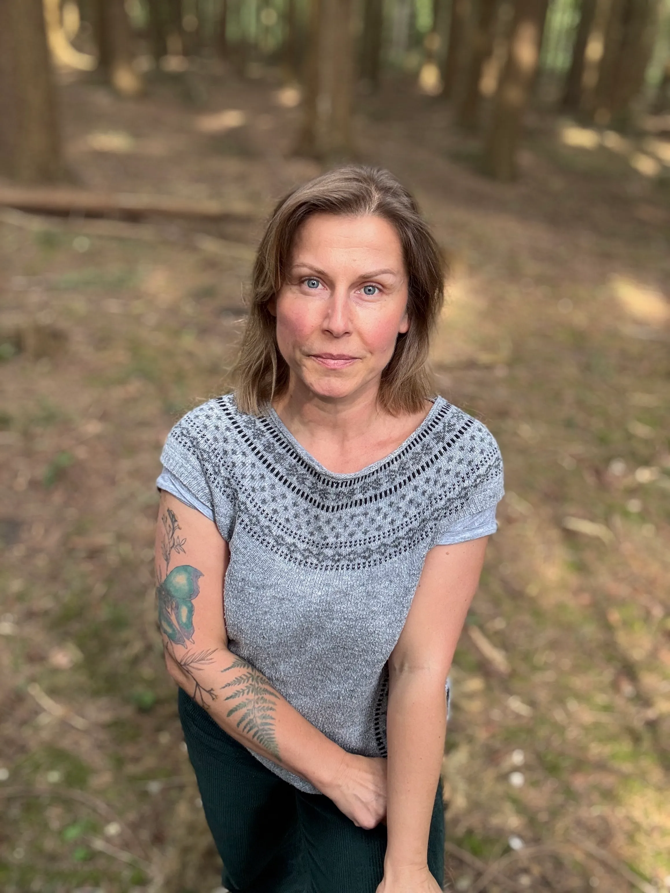 A woman with medium-length brown hair sits in a forest, looking directly at the camera. She has a thoughtful expression and is wearing a gray top with a decorative pattern. Her left arm is raised, resting on her shoulder, and is adorned with colorful tattoos, including a green bird and fern leaves. The background features tall trees and soft, dappled sunlight filtering through the foliage, creating a serene and natural atmosphere.